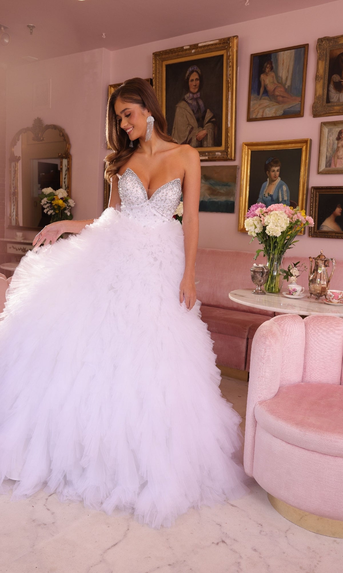 A woman wears the Ava Presley 29528 ruffled strapless ball gown with a beaded bodice, standing in a pink room adorned with vintage portraits, flowers, and a tea set on a round table.